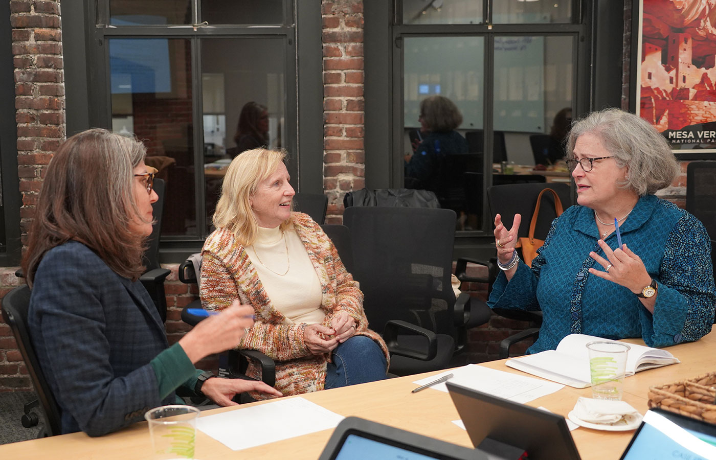 three-white-women-with-grey-hair-talk-enthusiastically-behind-a-table-with-papers-and-computers-on-it